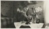 En glad kvinna (Ruth Svensson ?) sitter med en katt på axeln i ett sovrum.
Suddigt foto.