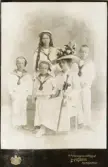 En kvinna i vit sommarklänning och stor hatt poserar tillsammans med sina fyra barn i ljusa sjömanskostymer m.m., hos fotografen.
Helfigur. Ateljéfoto.