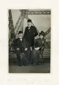 Två män i kostym och hatt m.m. och en kvinna i promenaddräkt poserar på en av avsatserna i Eiffeltornet, Paris.
Under fotot text på franska: 